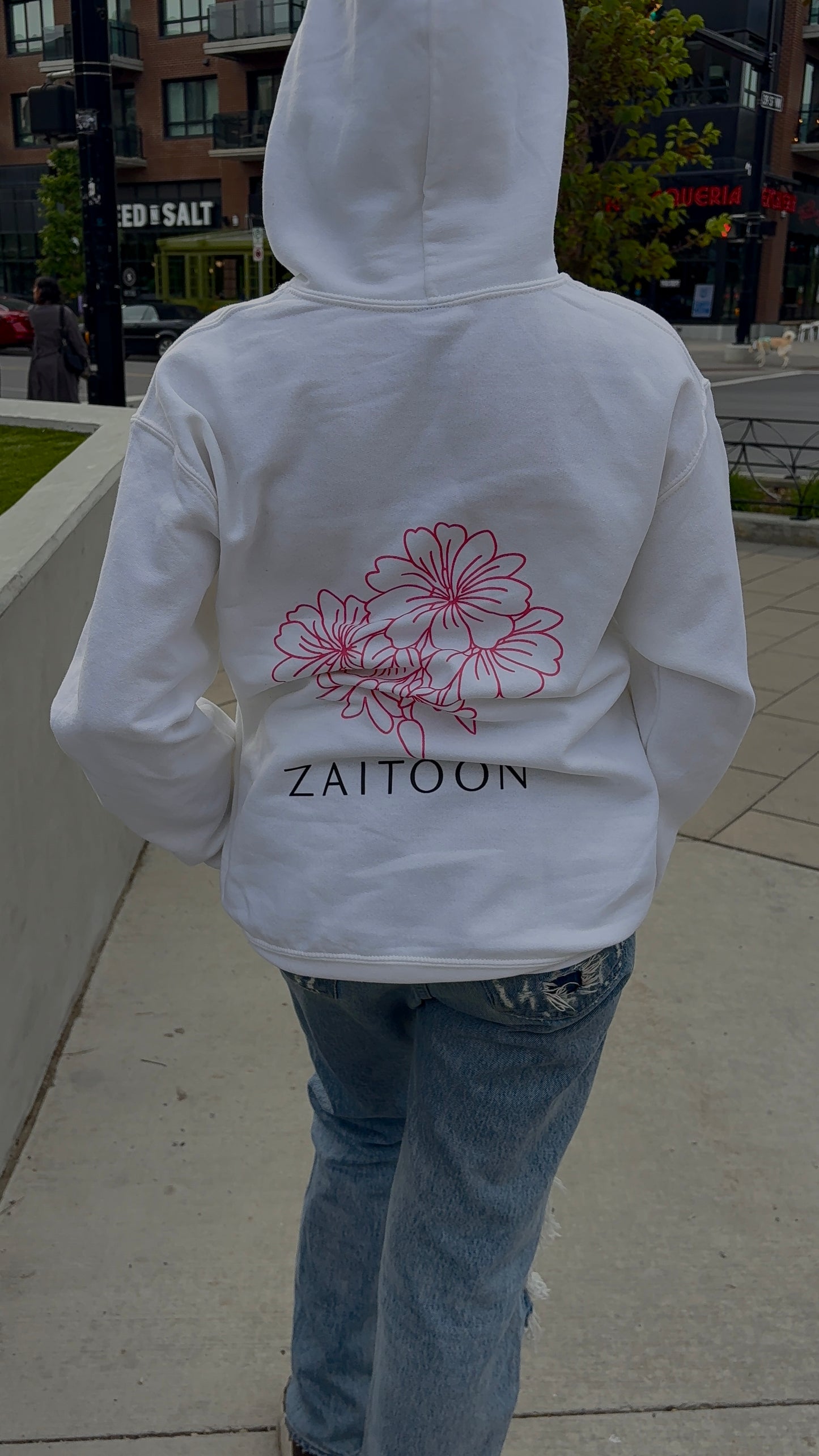 Person wearing a white hoodie with floral design and 'Zaitoon' text on the back, walking outdoors.
