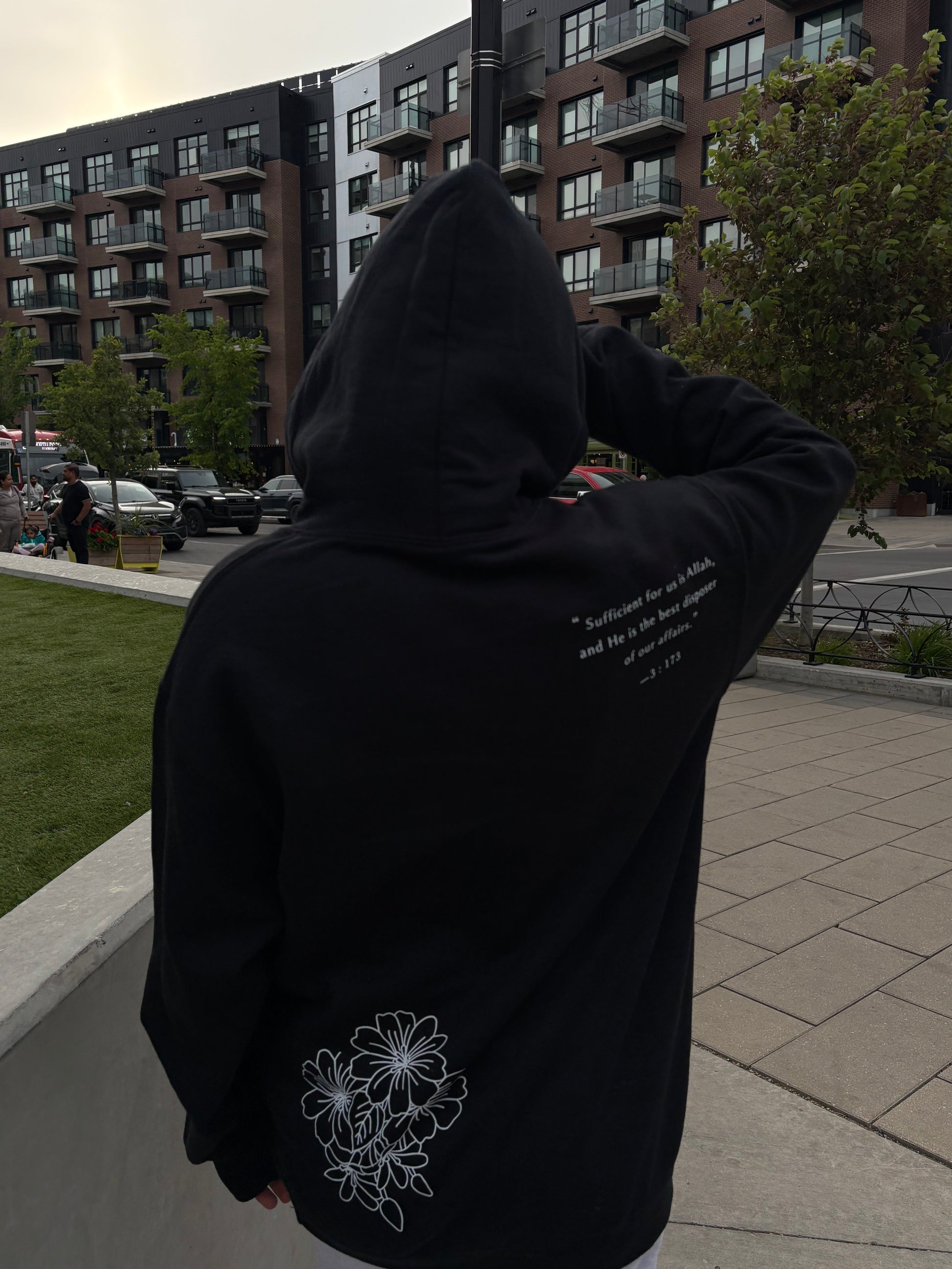 Person wearing a black hoodie with white text and floral design, standing on a sidewalk with buildings in the background.