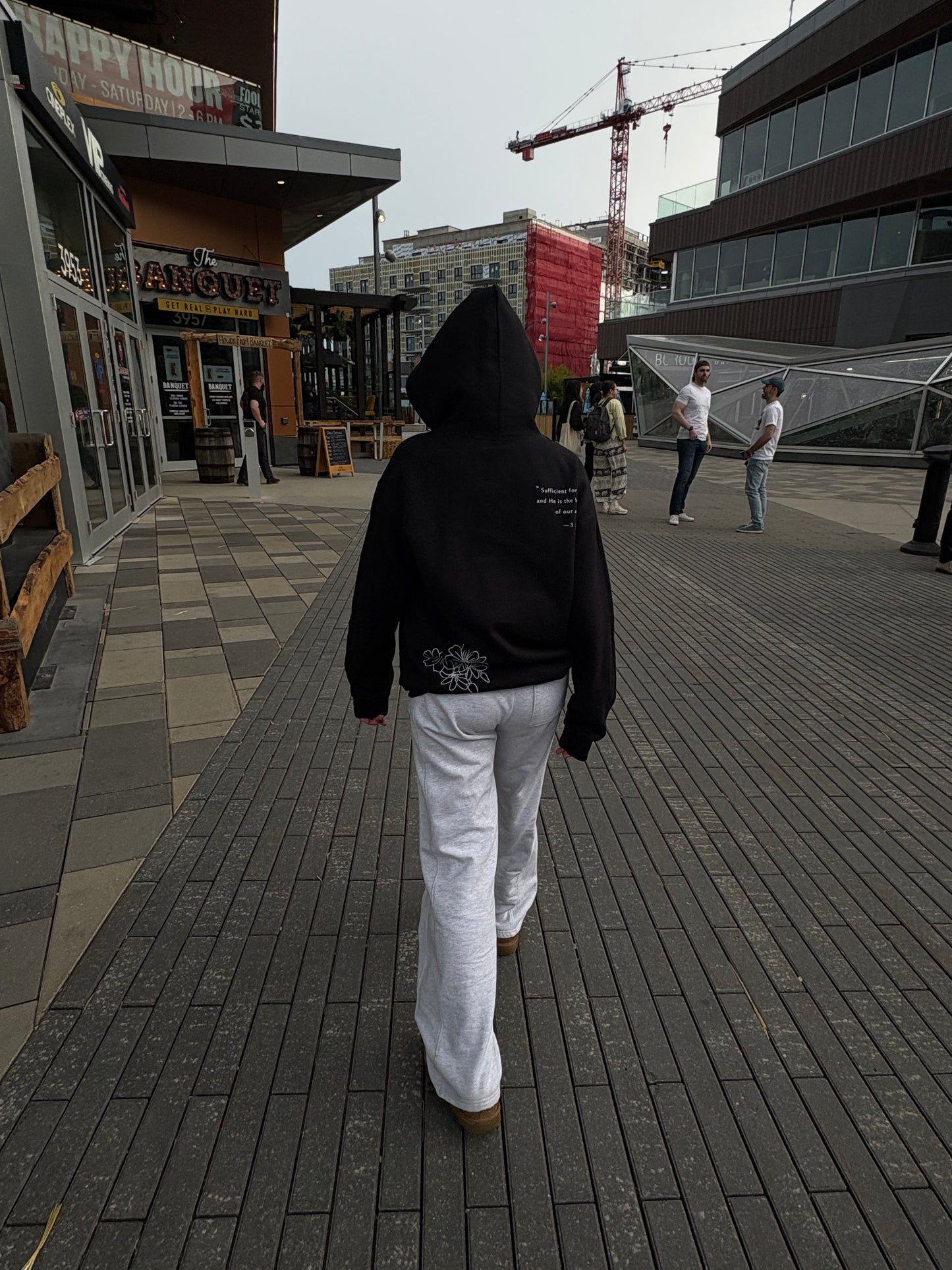 Person wearing a black hoodie and white pants walking on a sidewalk with buildings and cranes in the background.