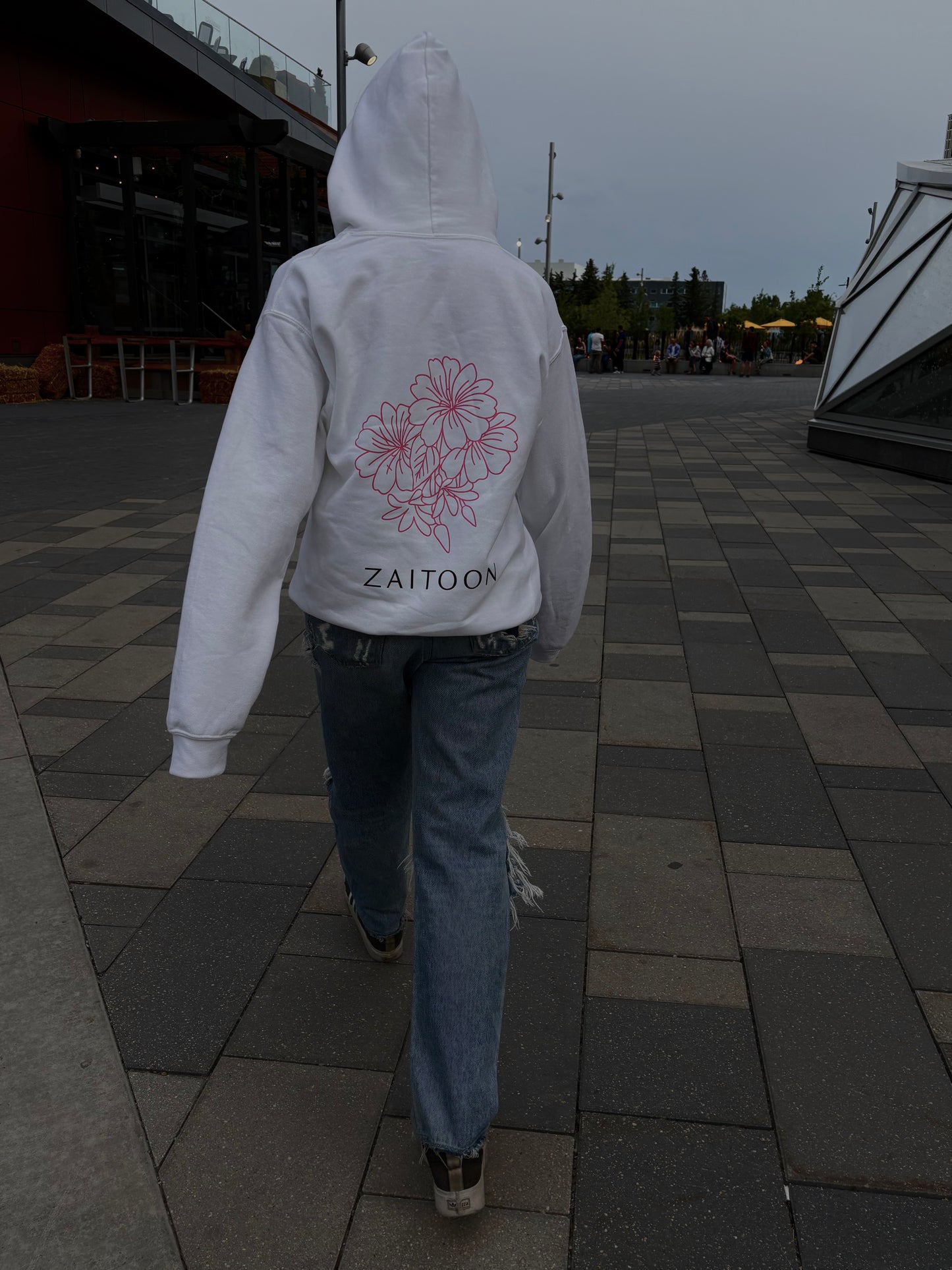Person wearing a white hoodie with floral design and 'Zaitoon' text, walking on a paved path.