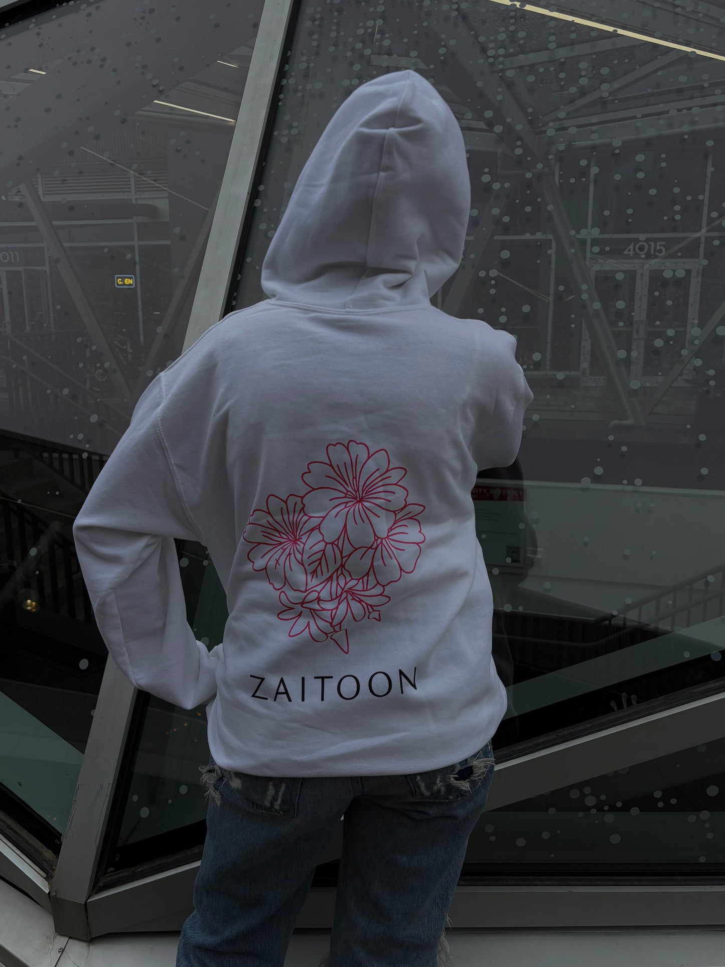 Person wearing a white hoodie with floral design and 'Zaitoon' text, standing in front of a large glass window.