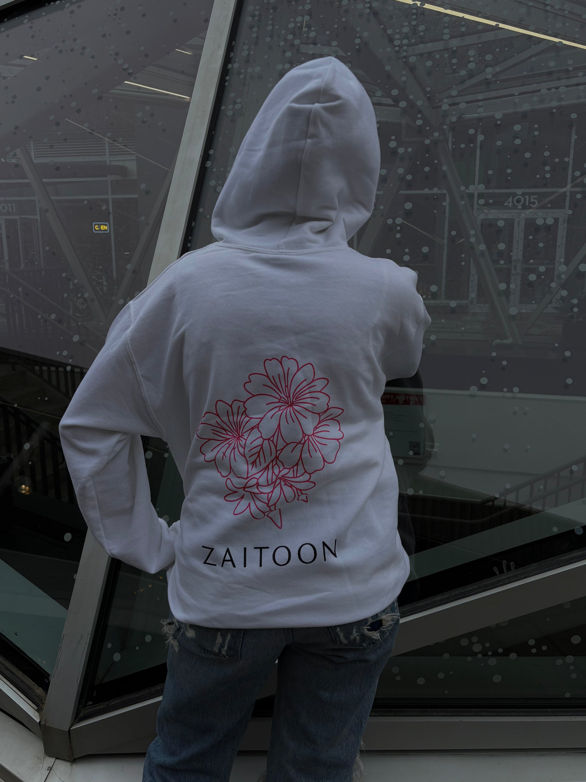 Person wearing a white hoodie with floral design and 'Zaitoon' text, standing in front of a large glass window.