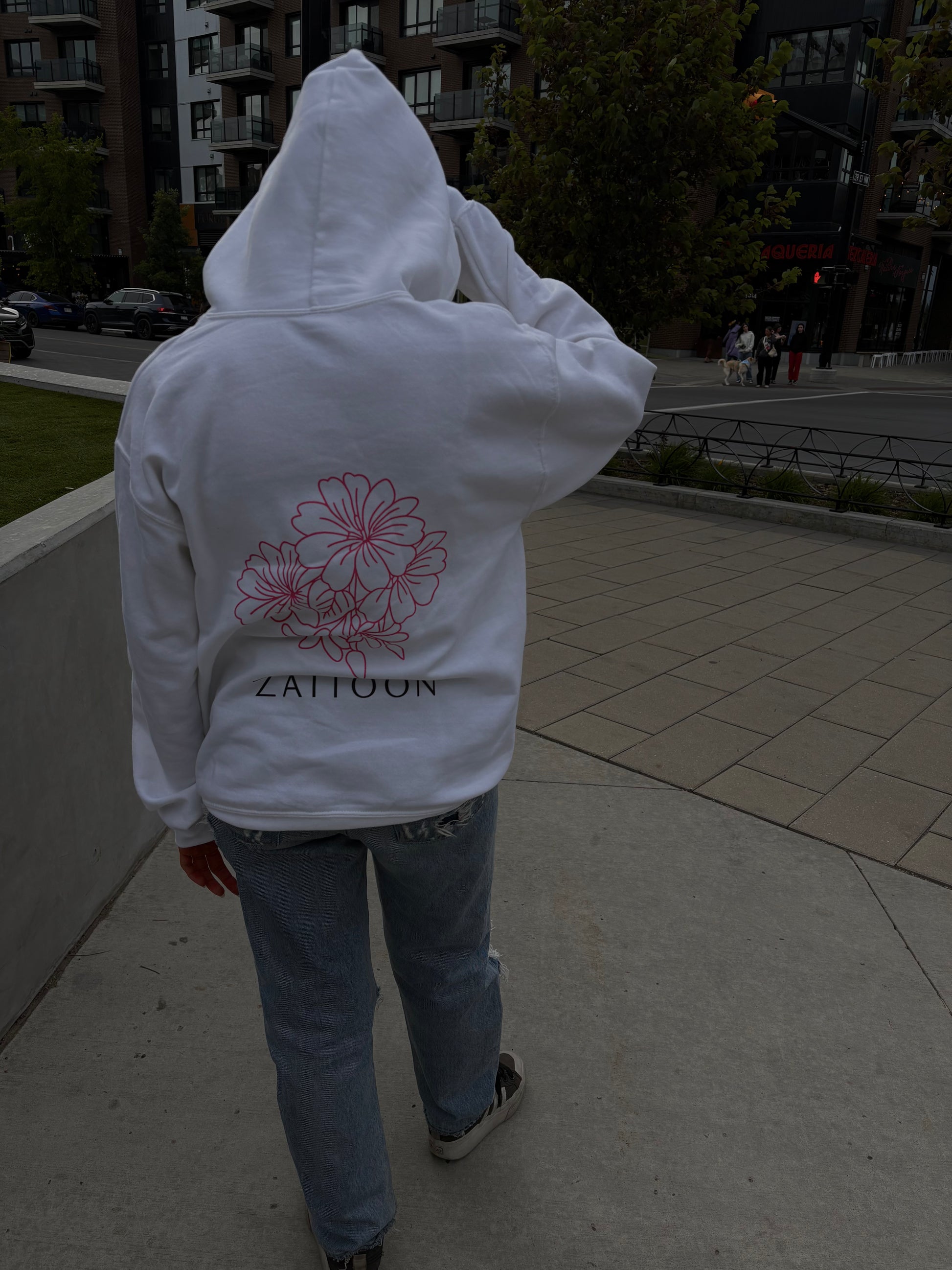 Person wearing a white hoodie with a floral design and 'Zattoon' text, walking on a sidewalk.